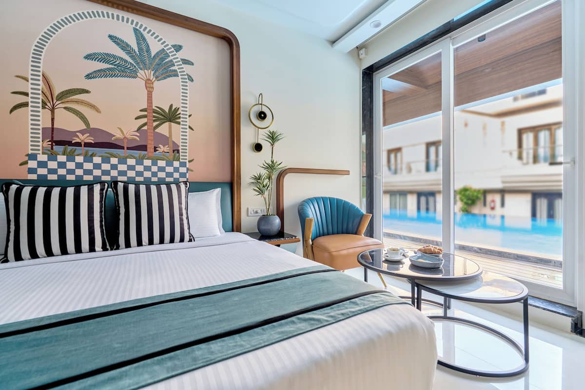 A hotel room with a large bed, a view of a swimming pool, a small round table, and a blue chair.