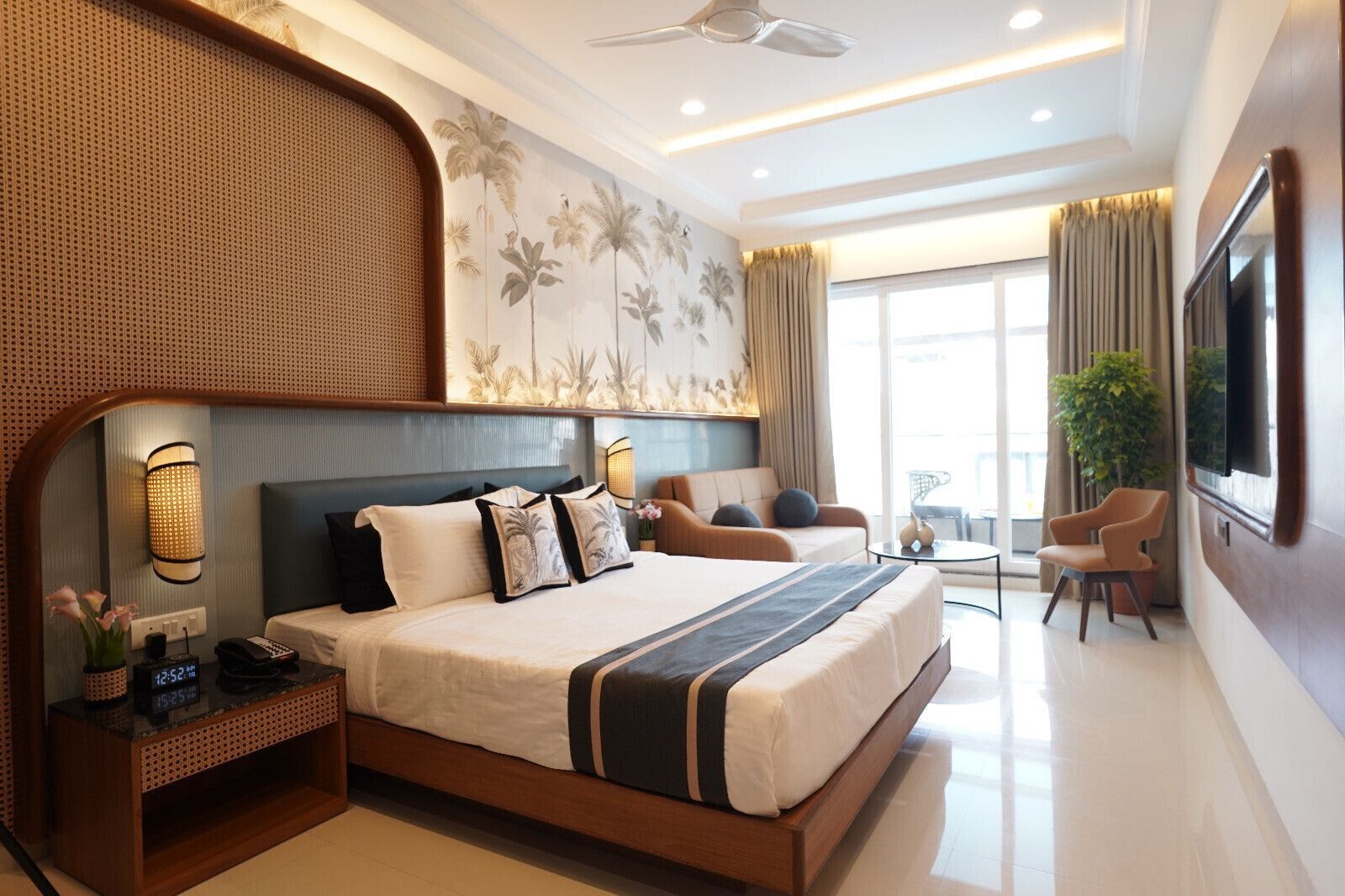 A modern hotel room with a large bed, a seating area with two chairs, a TV, and a large window with curtains.