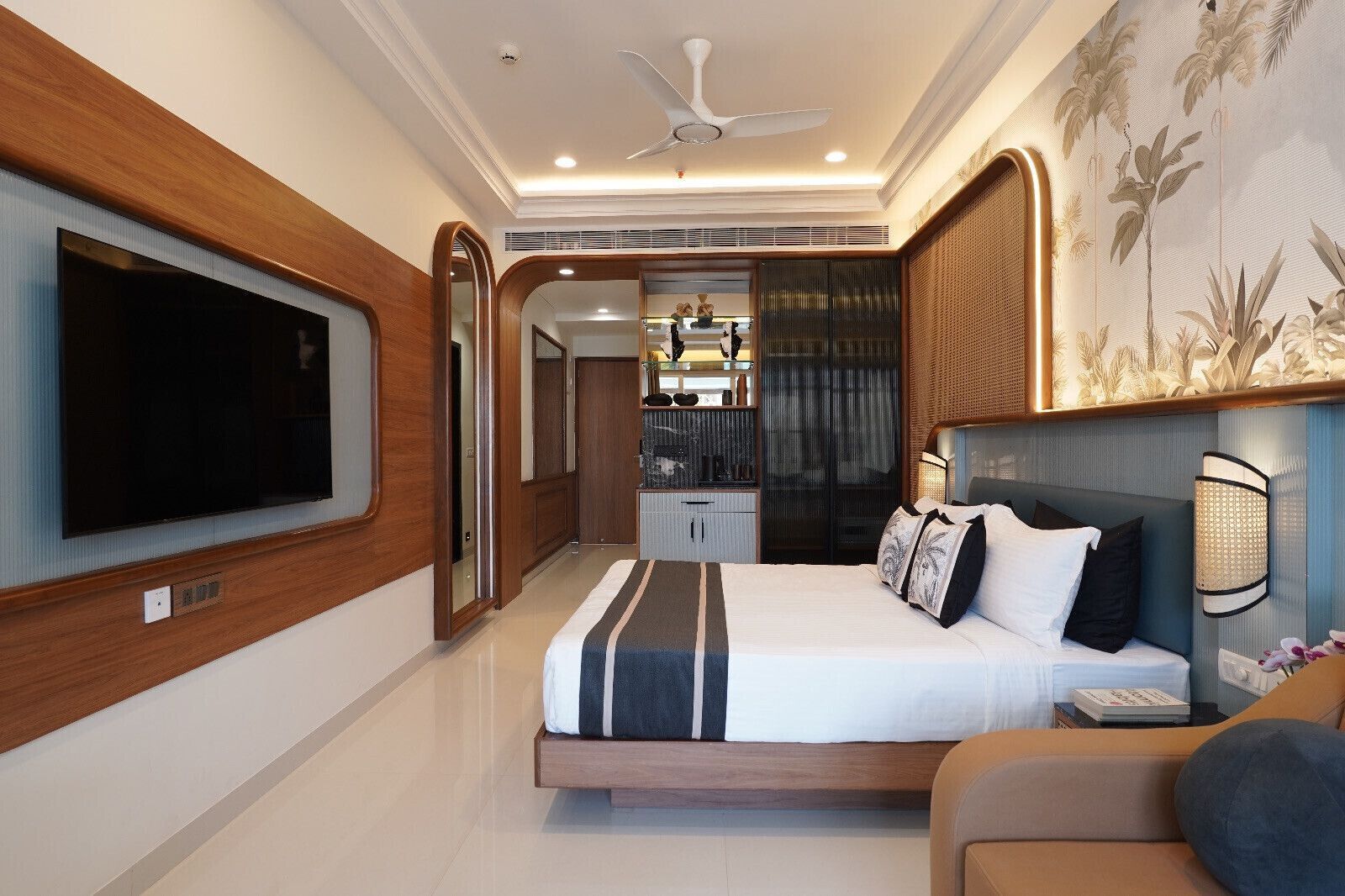A modern hotel room with a large flat-screen TV, a bed with a striped bedspread, a ceiling fan, and a decorative wall panel.