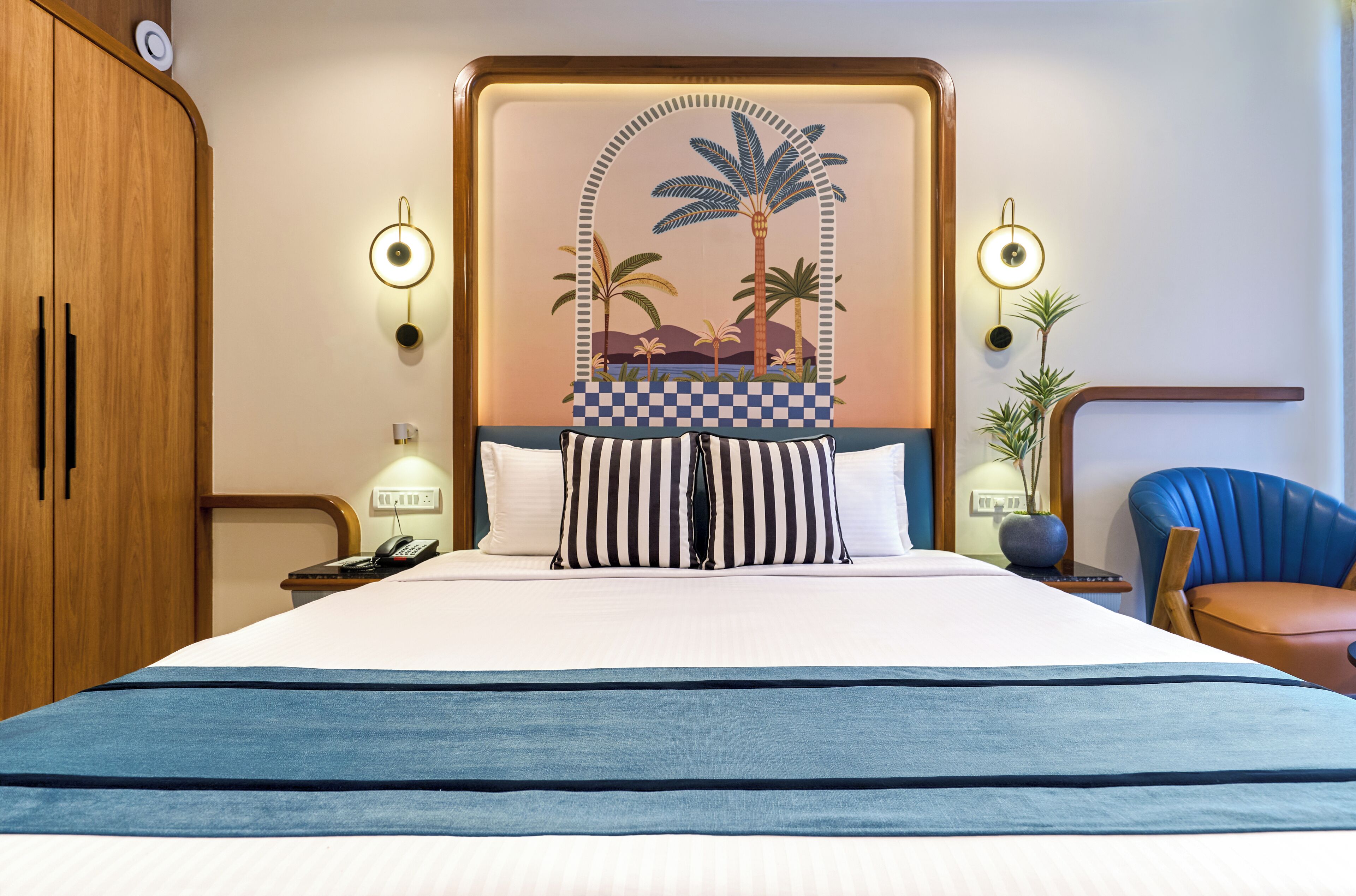 A hotel room with a large bed, a wooden wardrobe, a nightstand with a phone, two wall-mounted lamps, and a blue chair.