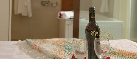 Una cama bien hecha con una bandeja que sostiene una botella y dos vasos, una bata colgada en un perchero y una zona de ducha al fondo.