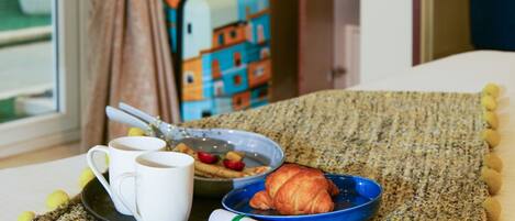 Habitación de hotel con cama, bandeja de desayuno, maleta y vistas al exterior.