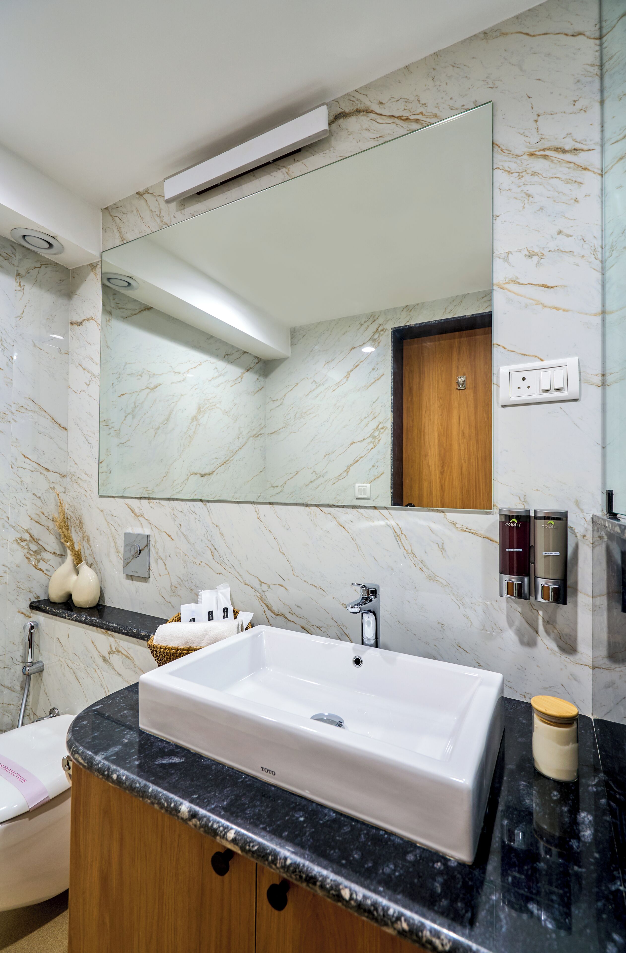 A modern bathroom with a large mirror, marble walls, a white sink, and a wooden cabinet.