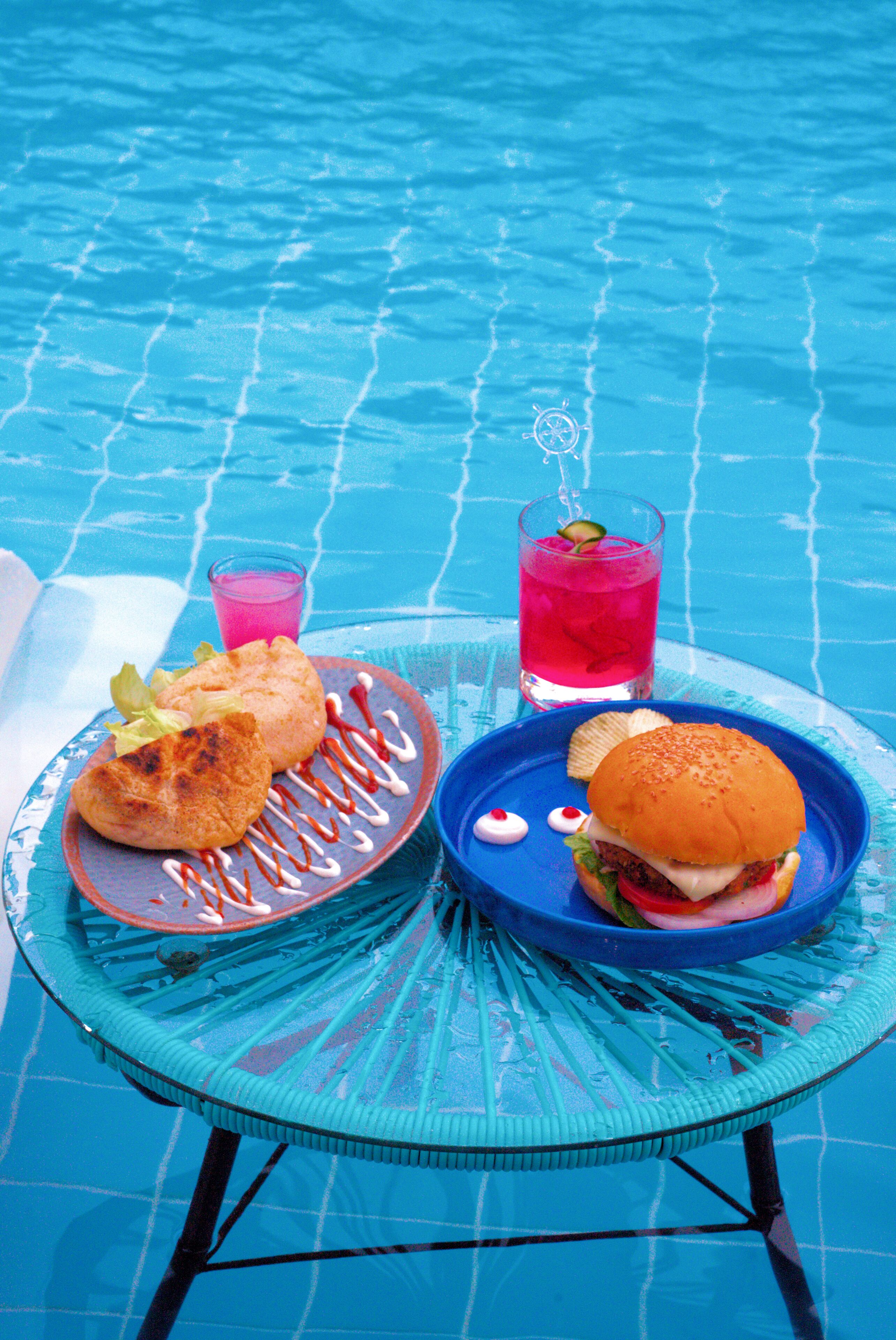 A table by a pool with a plate of food and drinks.
