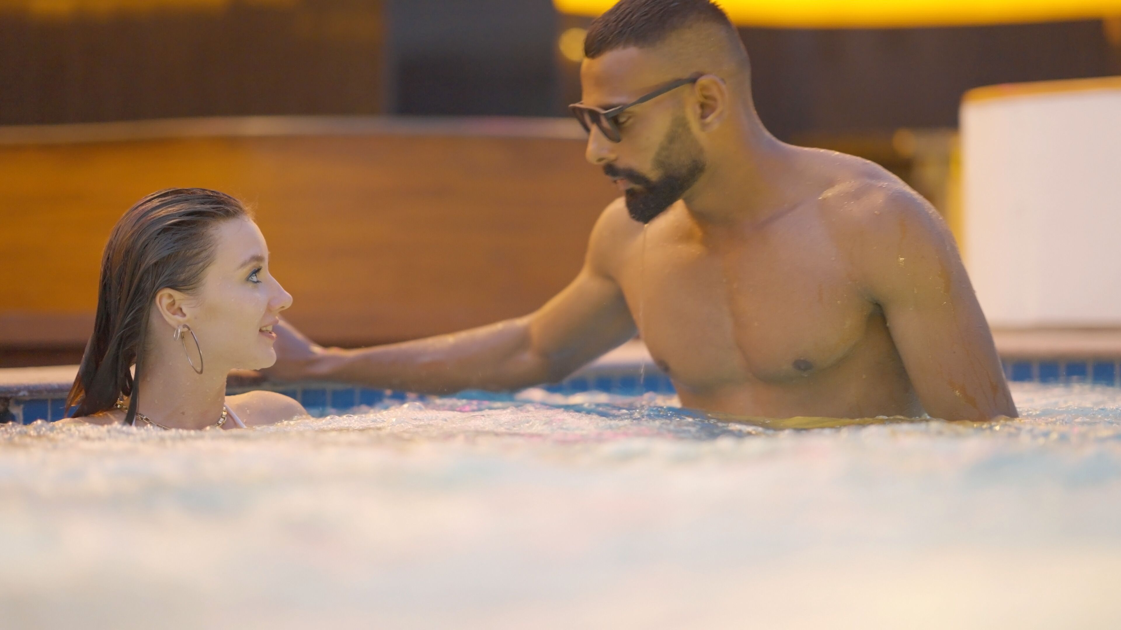 Un couple dans un jacuzzi, avec un sol carrelé et une structure en bois en arrière-plan.