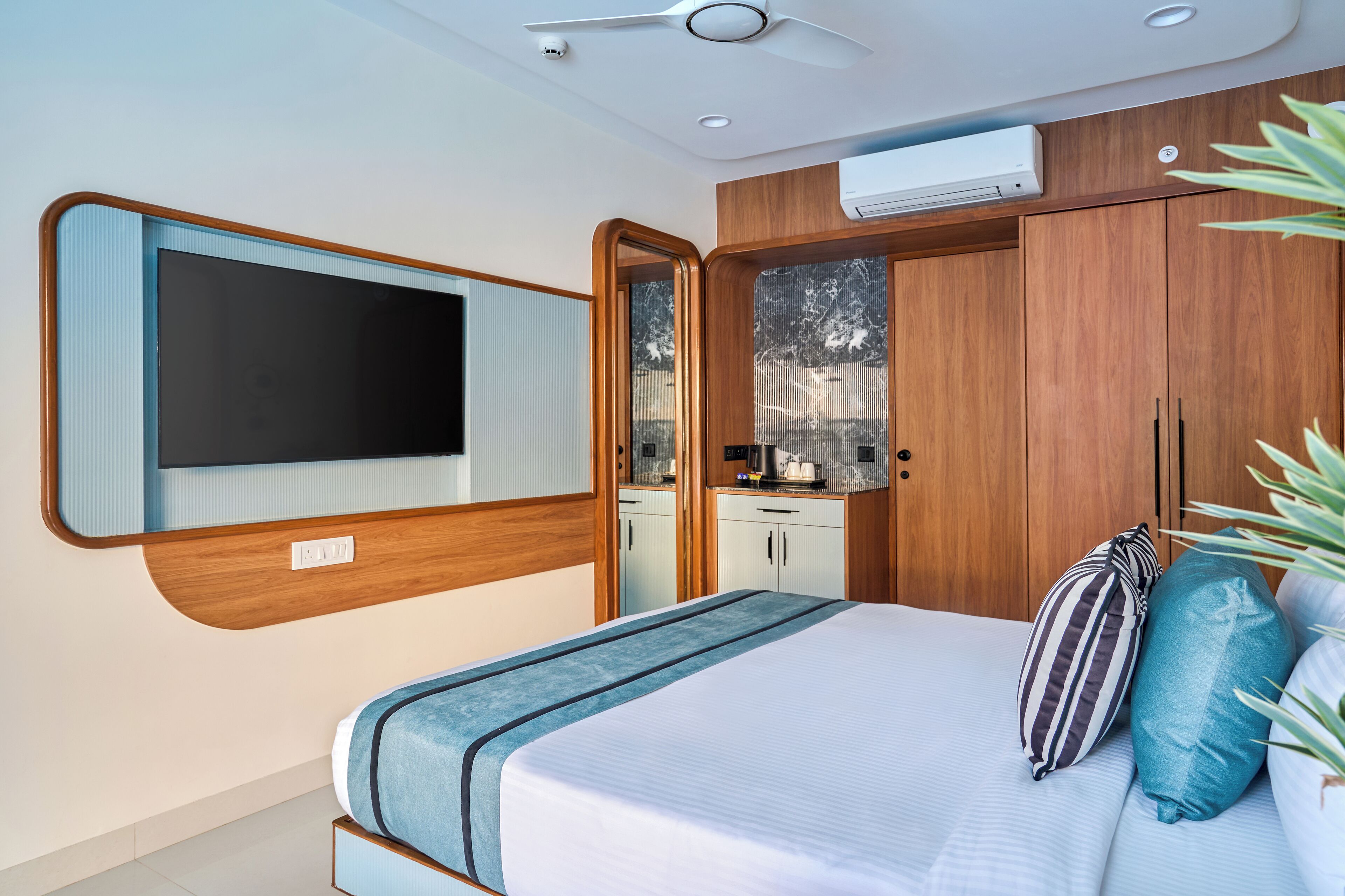 A modern bedroom with a bed, a TV mounted on the wall, wooden cabinets, and a small kitchenette.