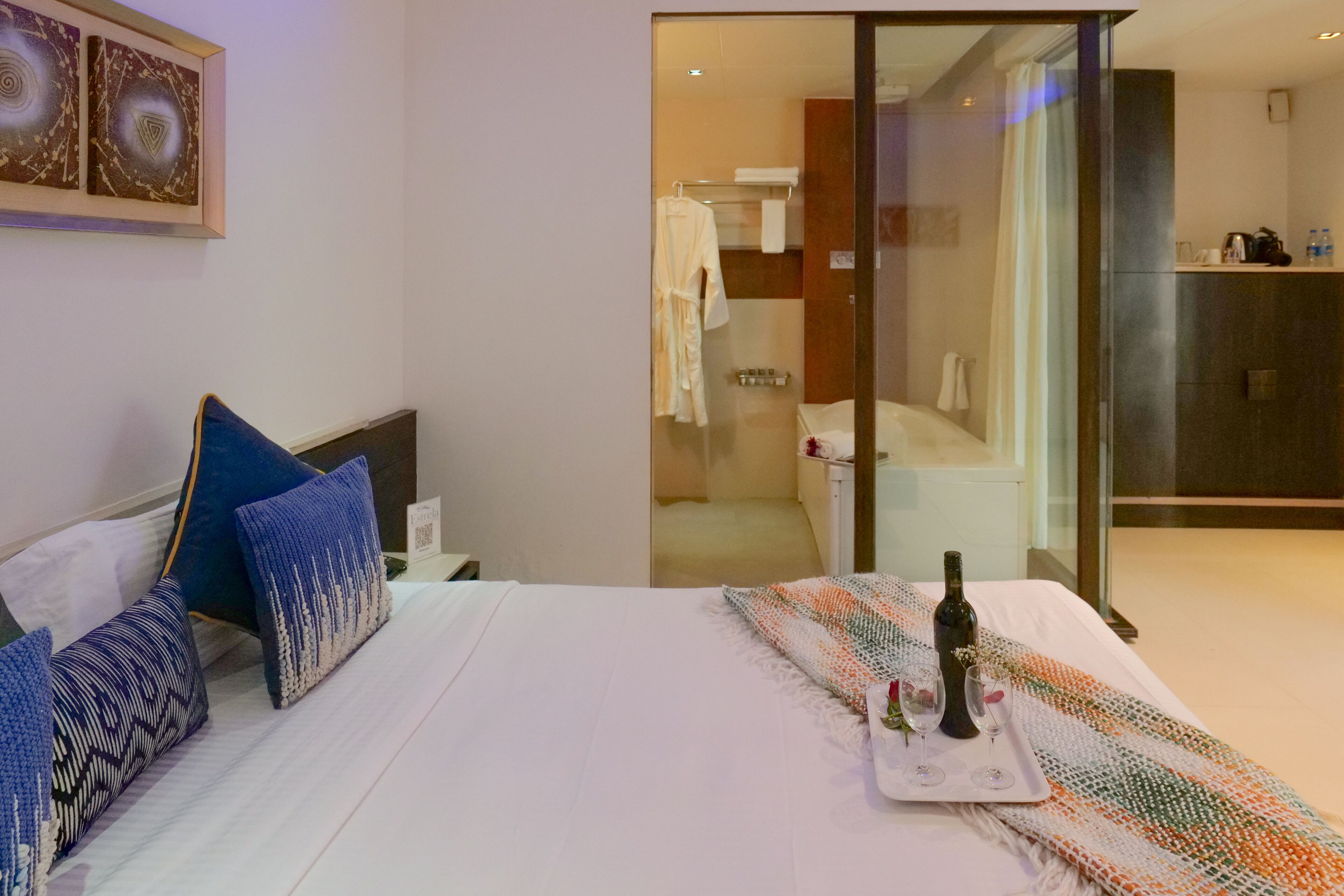 A hotel room with a bed, pillows, a tray with a bottle and glasses, and a view of the bathroom.
