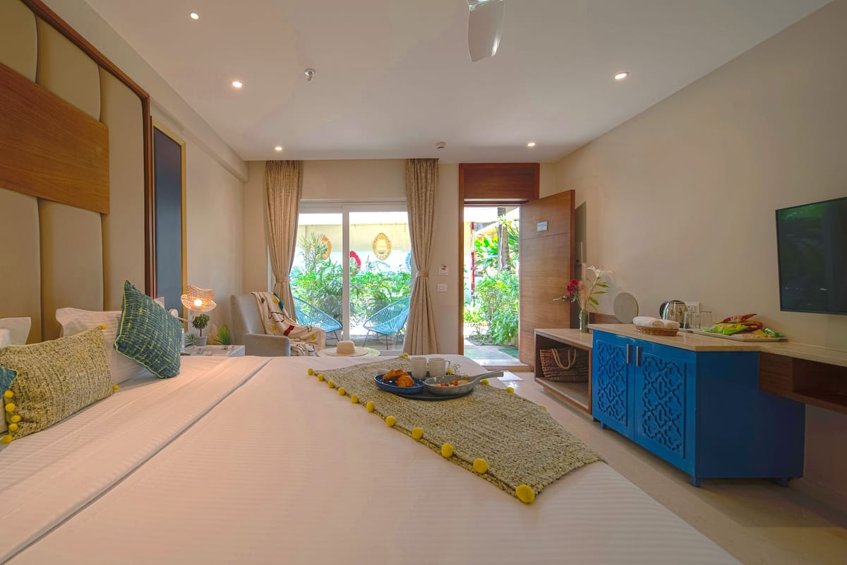 A hotel room with a large bed, a sitting area with a table, a TV, and a view of greenery outside.