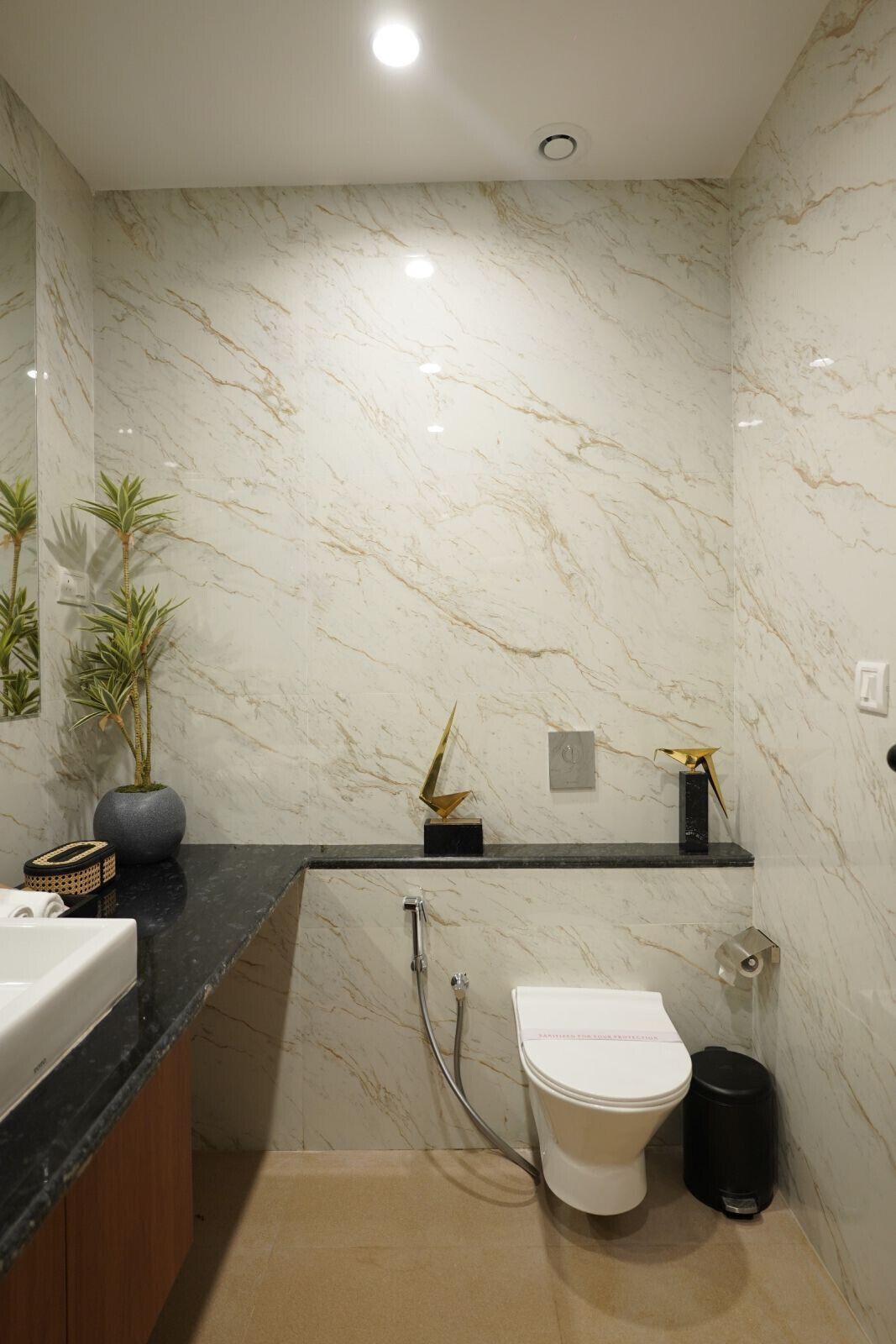 A modern bathroom with a marble wall, a white toilet, a black trash bin, and a small shelf with decorative items.