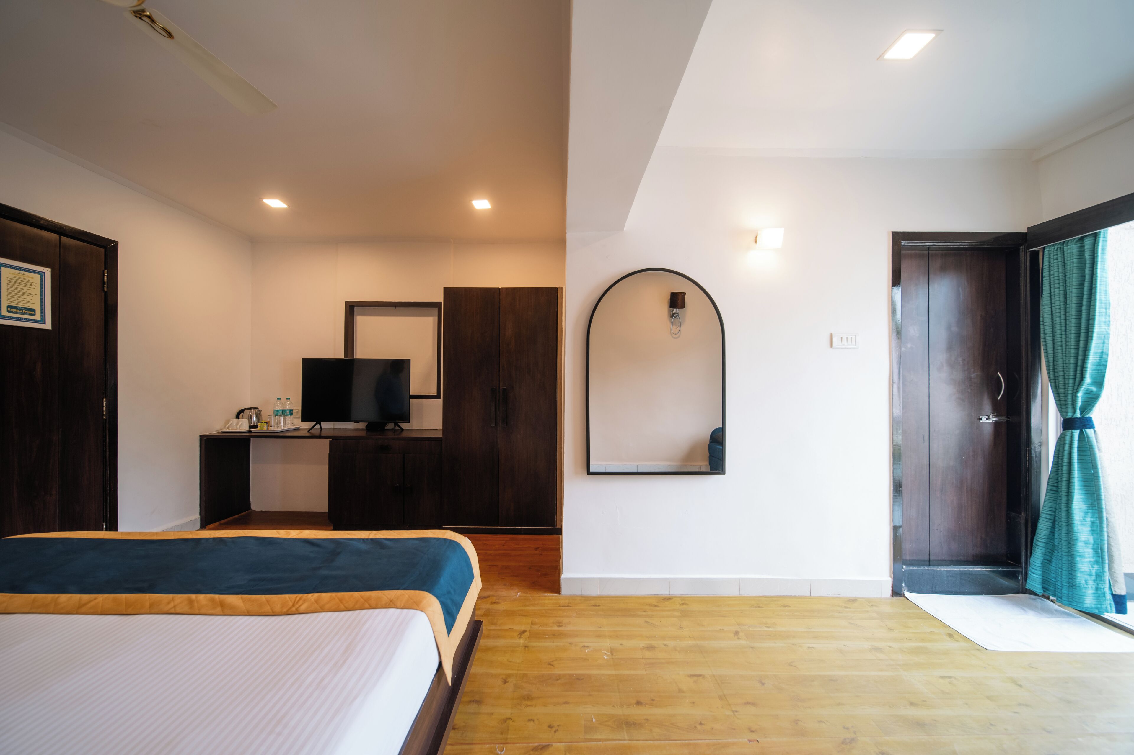 Ein Hotelzimmer mit einem Bett, einem Schreibtisch mit Fernseher, einem Spiegel und einer Tür mit Vorhängen.