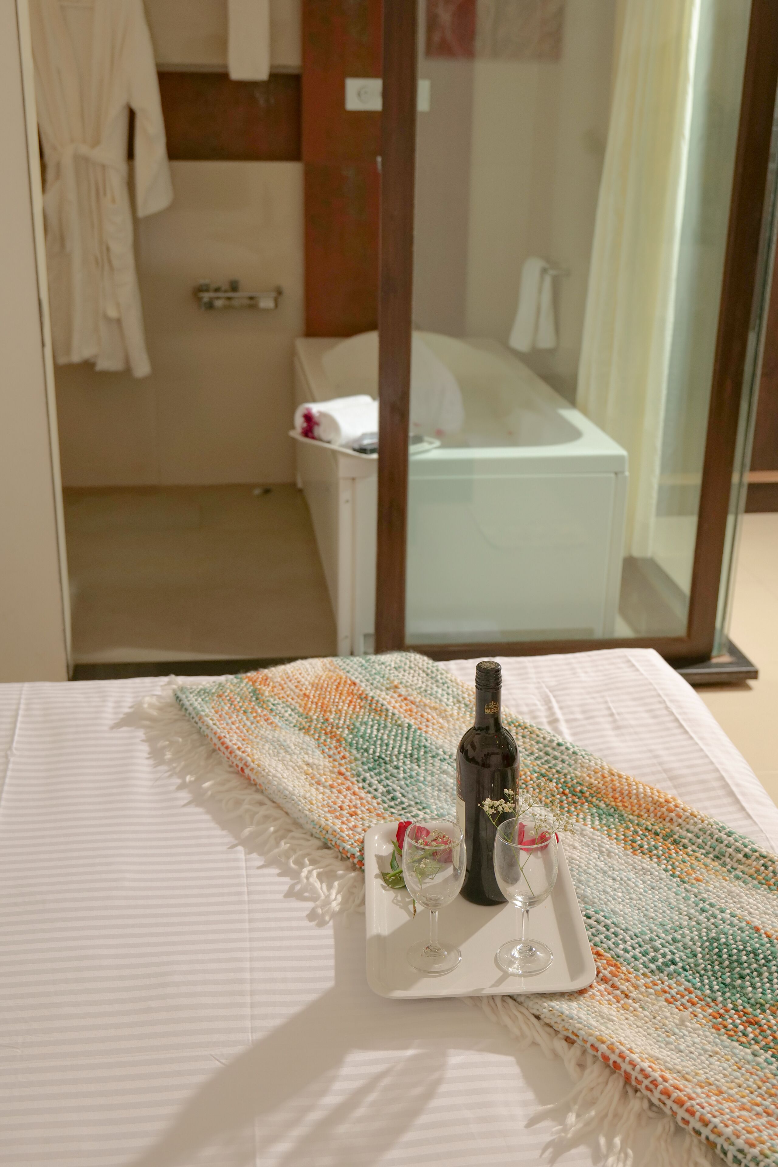 A hotel room with a bed, a glass shower enclosure, a white bathtub, and a tray with a bottle and glasses on the bed.