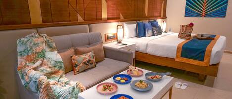 A hotel room with a sofa, a bed, a dining table with breakfast items, and a wall with a geometric pattern.