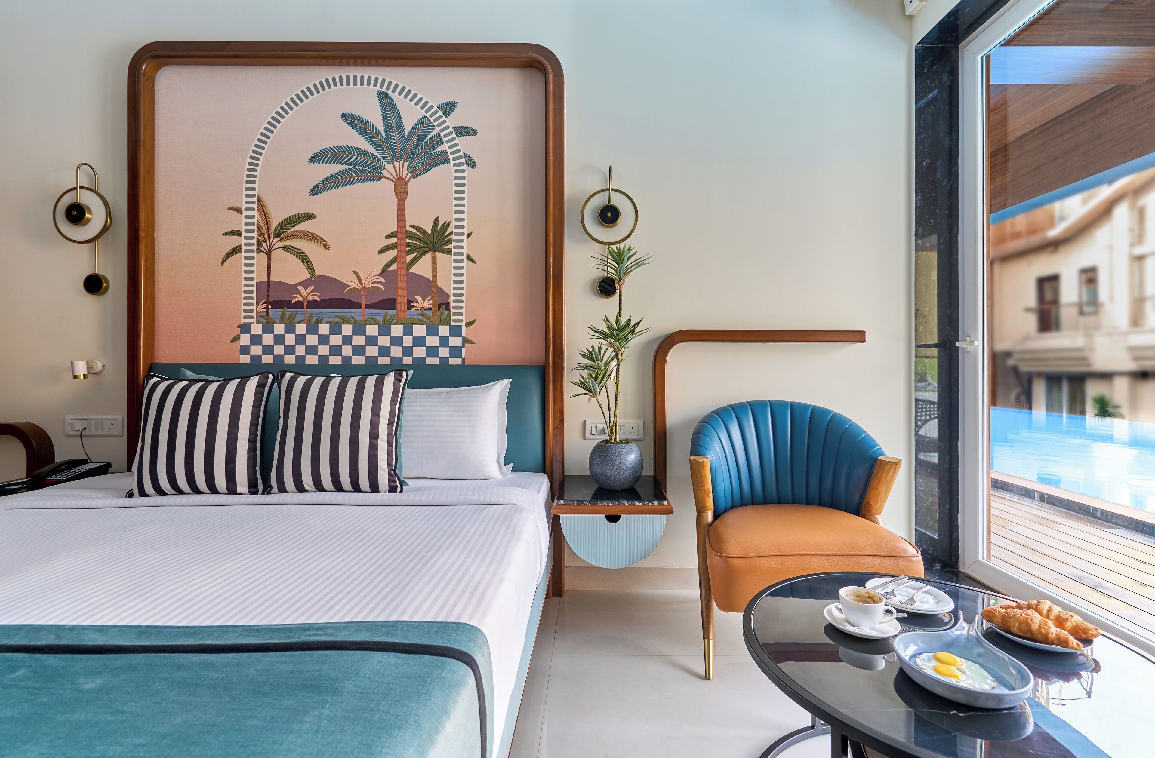 Une chambre d’hôtel avec un lit, un fauteuil bleu, une petite table avec un petit-déjeuner, et une vue sur une piscine.