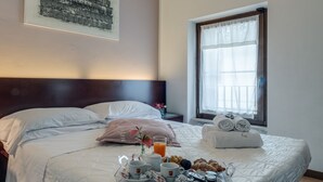 Una cama bien hecha con sábanas blancas, una bandeja de desayuno con croissants, bollería y café, y una ventana con vistas al exterior.