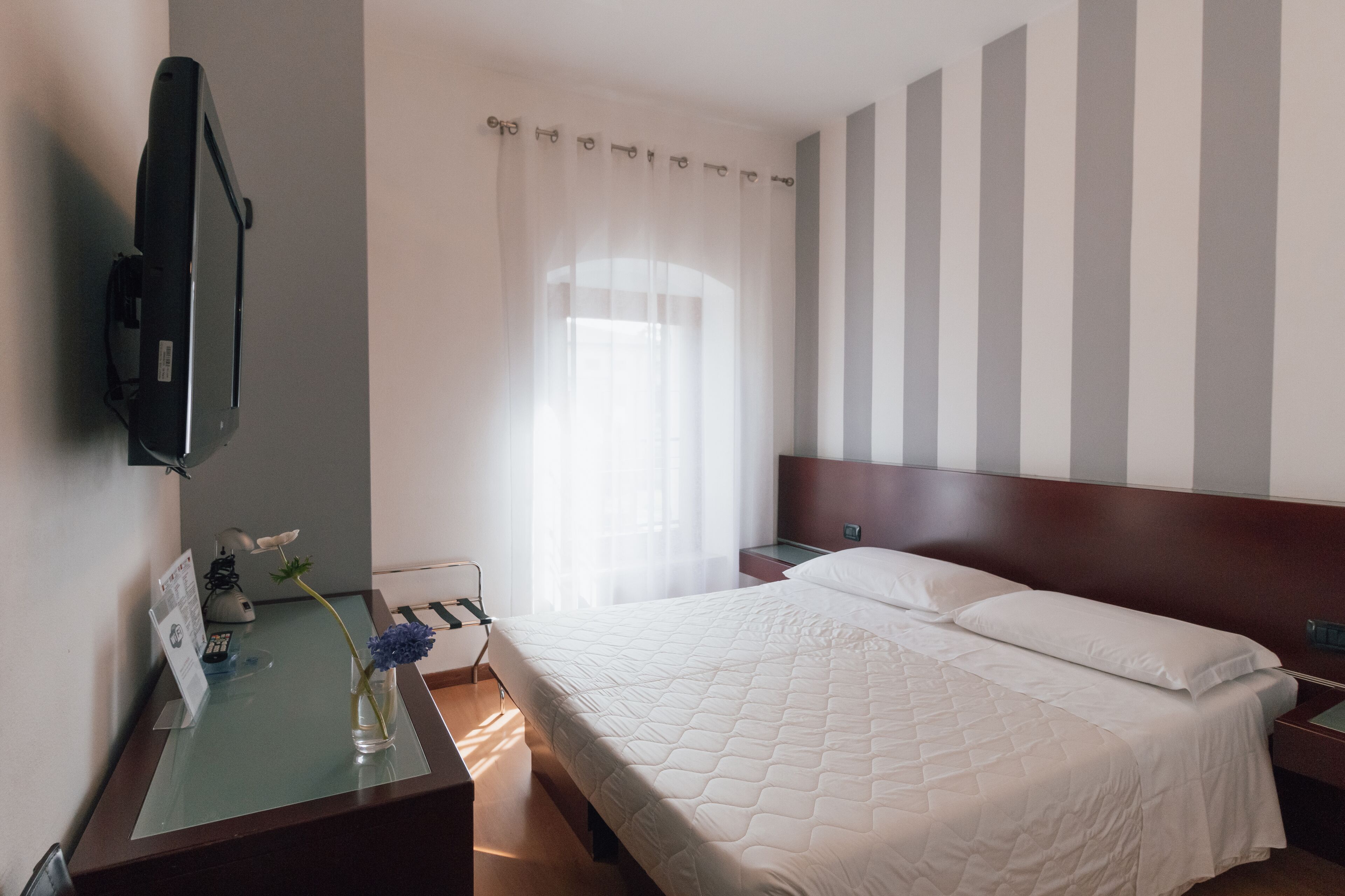 A hotel room with a bed, a desk with a computer, a TV mounted on the wall, and a window with curtains.