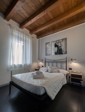 A bedroom with a wooden ceiling, a bed with white bedding, two bedside tables with lamps, and a window with sheer curtains.