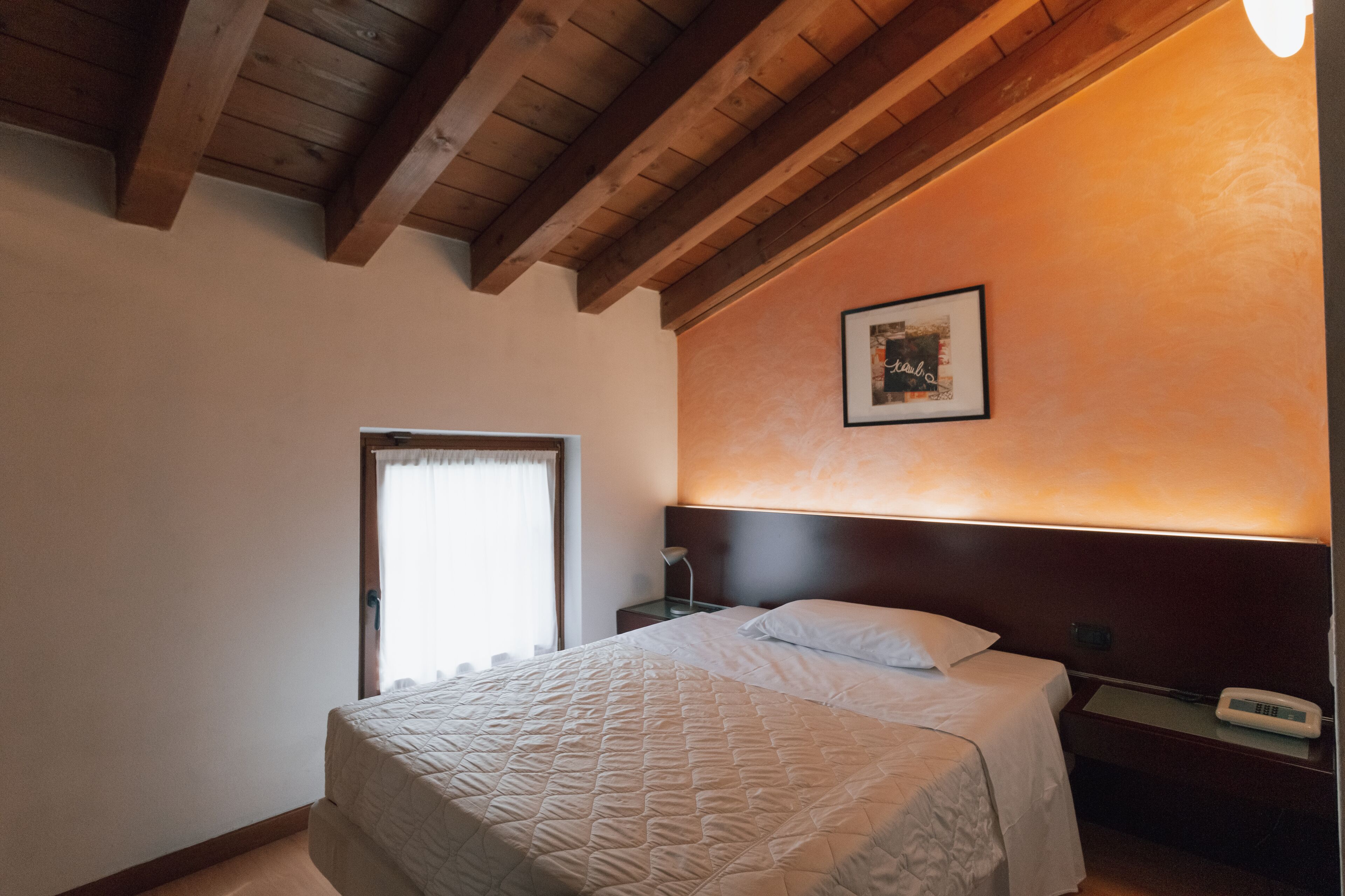 Una habitación de hotel con cama, mesita de noche, un cuadro enmarcado en la pared y una ventana con cortinas.