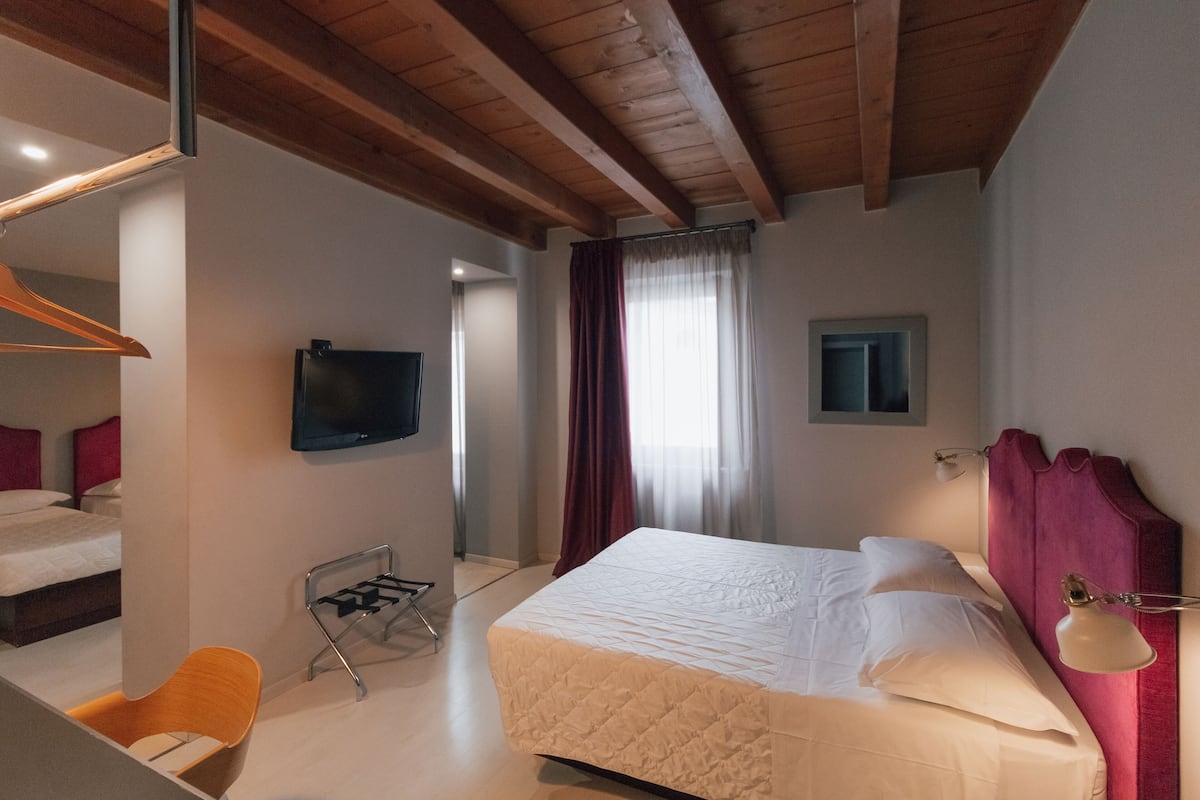 Ett hotellrum med en säng, en tv som är monterad på väggen, ett skrivbord med en stol och ett fönster med gardiner.
