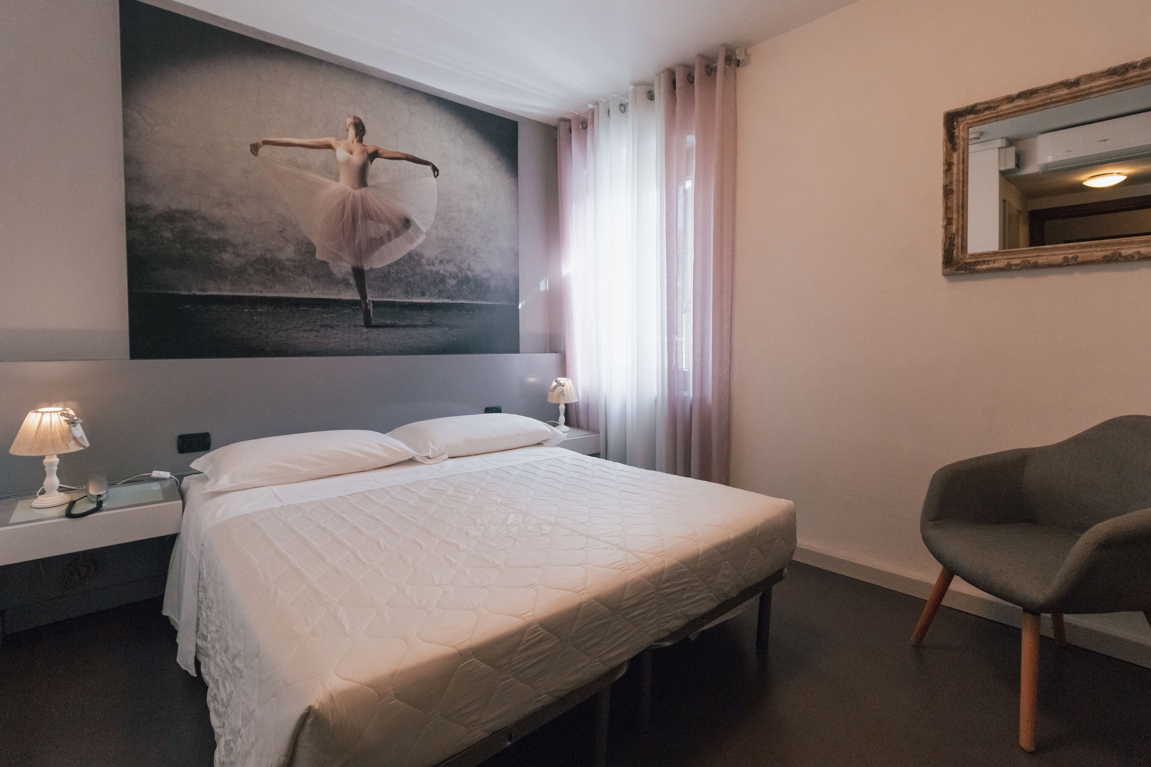 A hotel room with a large bed, a nightstand with a lamp, a chair, a wall-mounted mirror, and a ballet dancer painting.