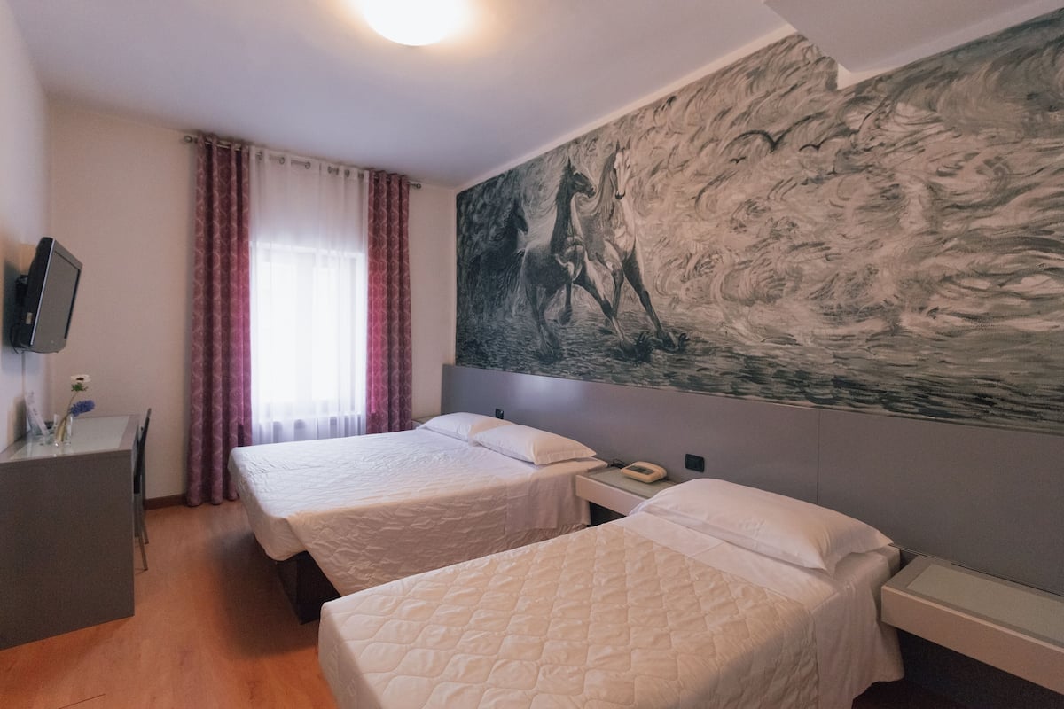 Ett hotellrum med två sängar, ett skrivbord, en stol, en tv på väggen och en stor tavla med hästar.