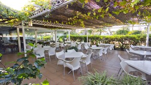 An outdoor cafe with white tables and chairs, surrounded by greenery and a covered area with a lattice structure. - Hotel Campomar Playa (El Puerto de Santa Maria)