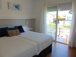 Cama doble con ropa de cama blanca y azul, una mesita de noche pequeña con lámpara, y vista a un balcón con mobiliario exterior.