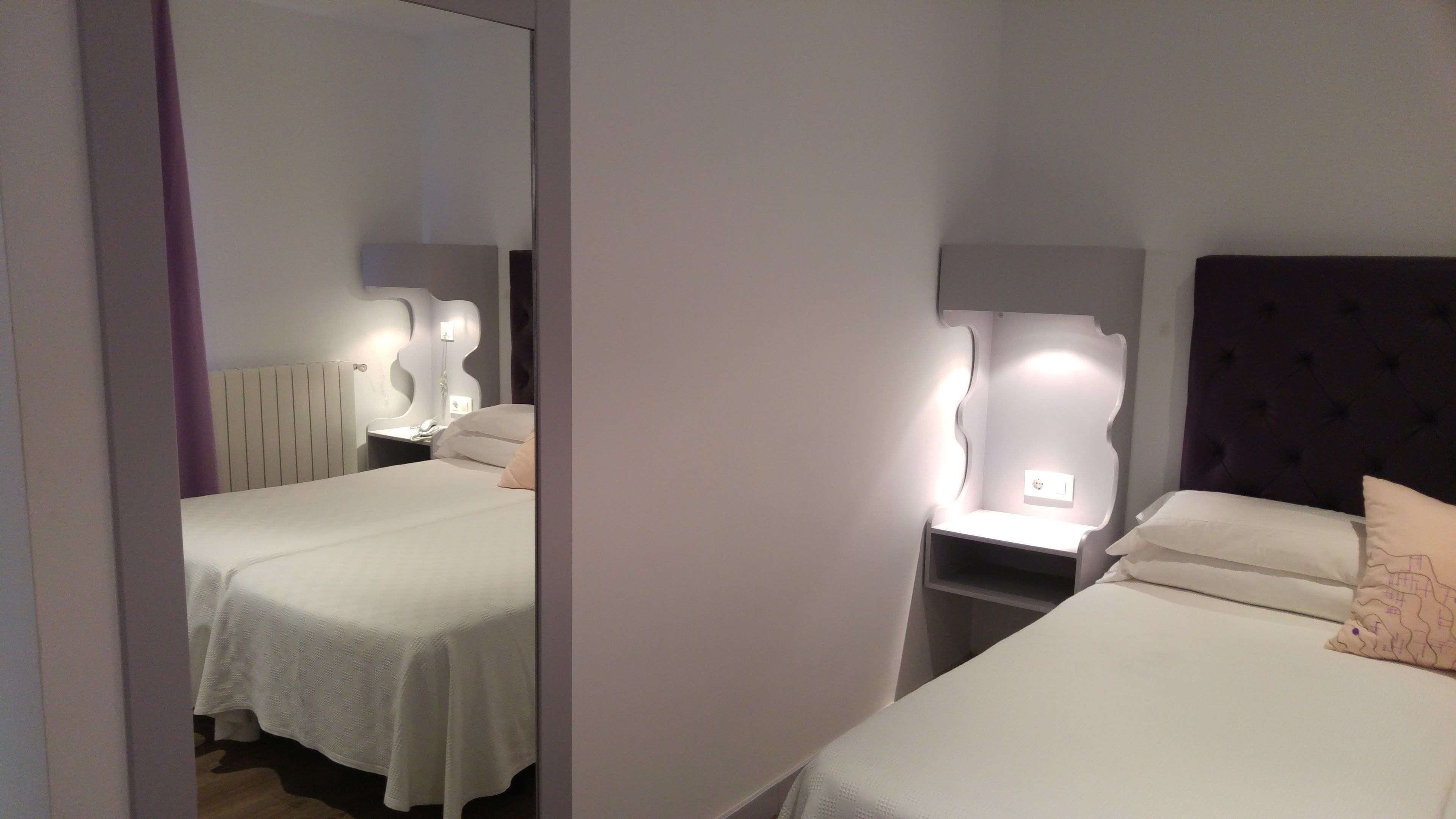 A hotel room with two beds, a mirror, bedside tables, and a wall-mounted light fixture.