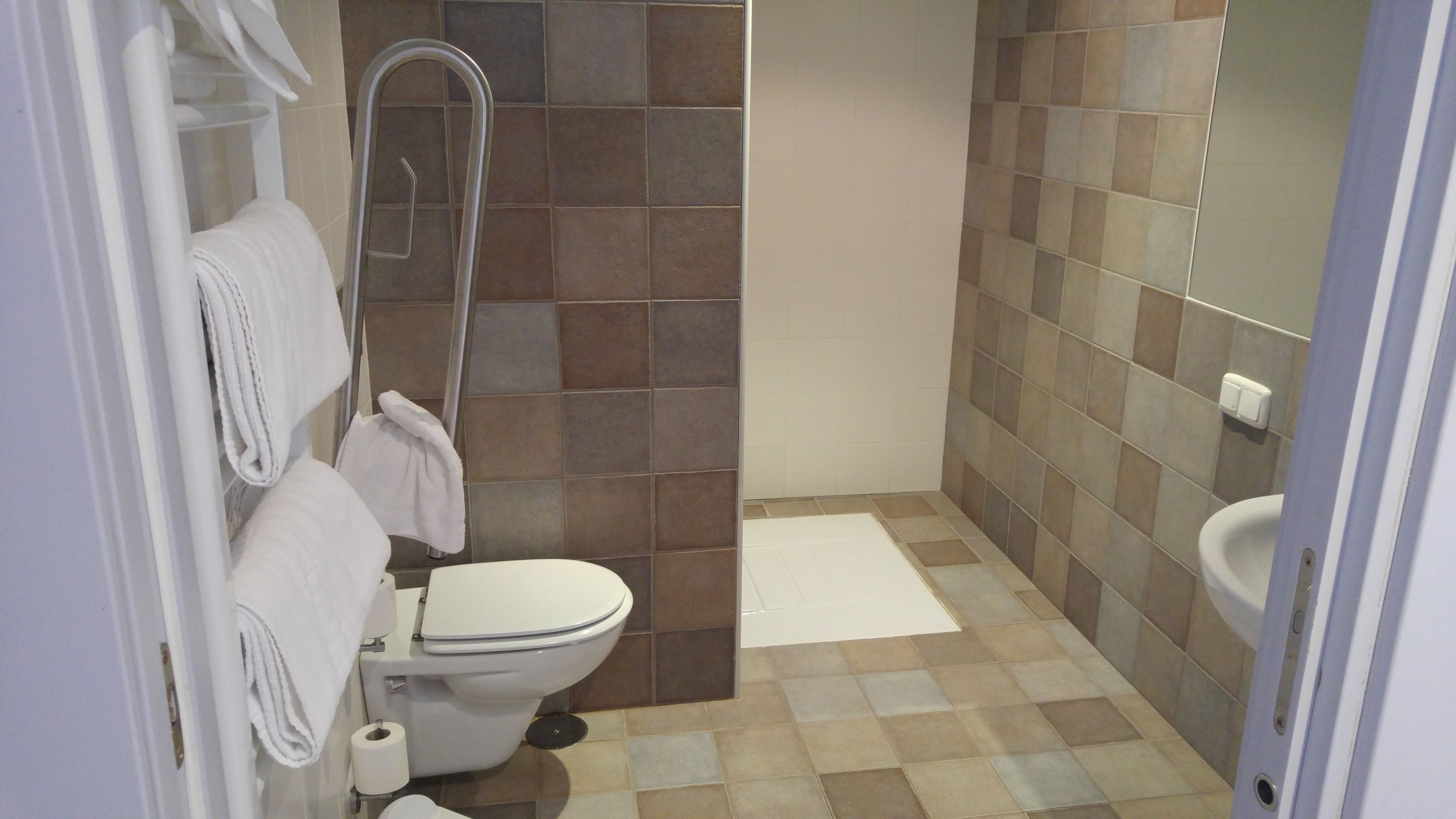 Een badkamer met toilet, douche en douchebrons. Handdoeken hangen aan de muur en er ligt een rol toiletpapier op de vloer.