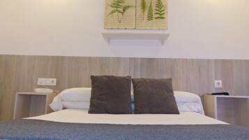 A neatly made bed with two pillows, a bedside table with a phone, and a wall-mounted shelf with botanical illustrations.