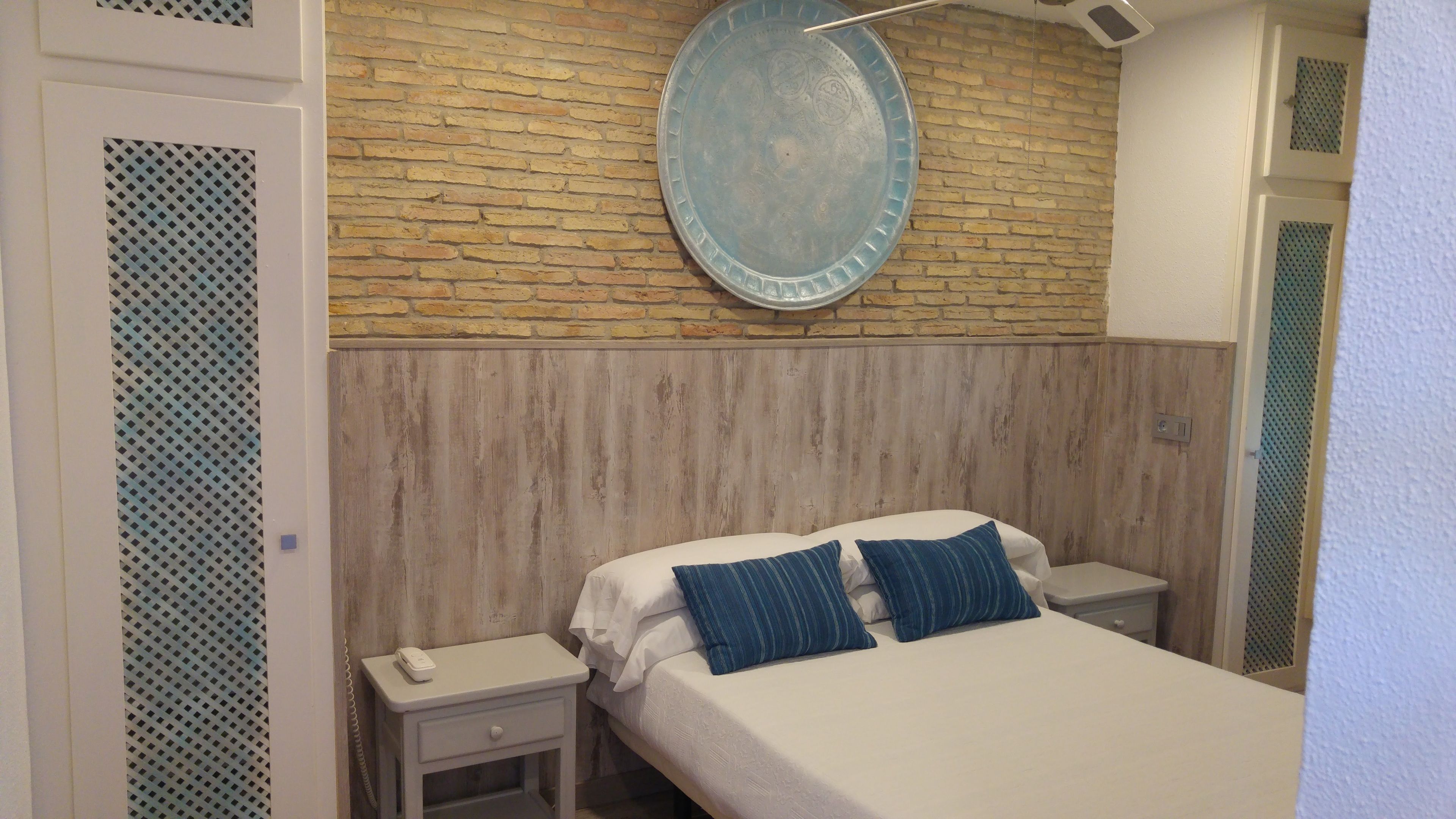 Quarto com cabeceira de madeira, cama com roupa de cama branca e dois travesseiros azuis, mesa de cabeceira com um telefone e um prato decorativo azul na parede.