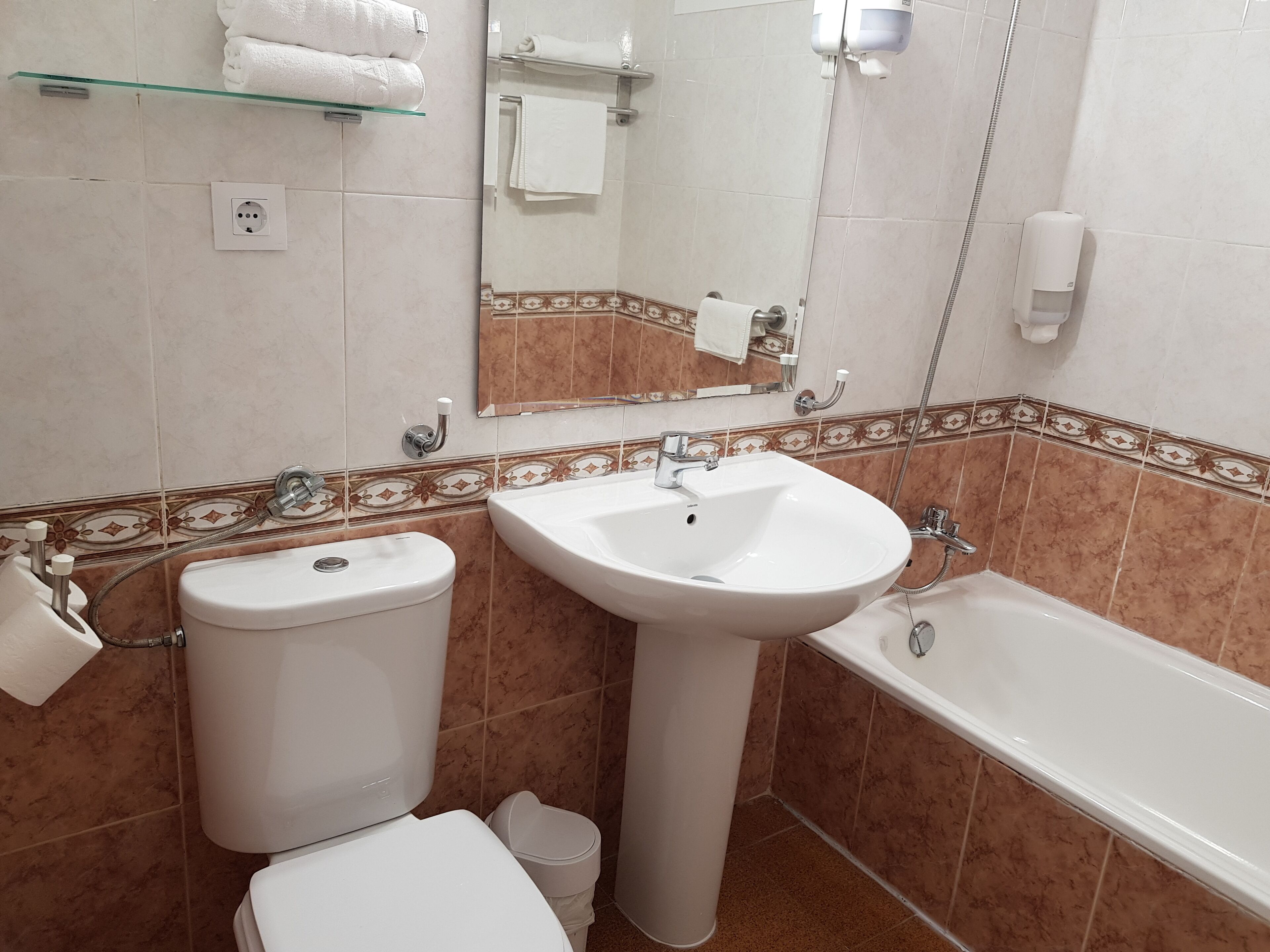 A bathroom with a toilet, sink, and bathtub. There are towels on a shelf, a mirror, and a soap dispenser.