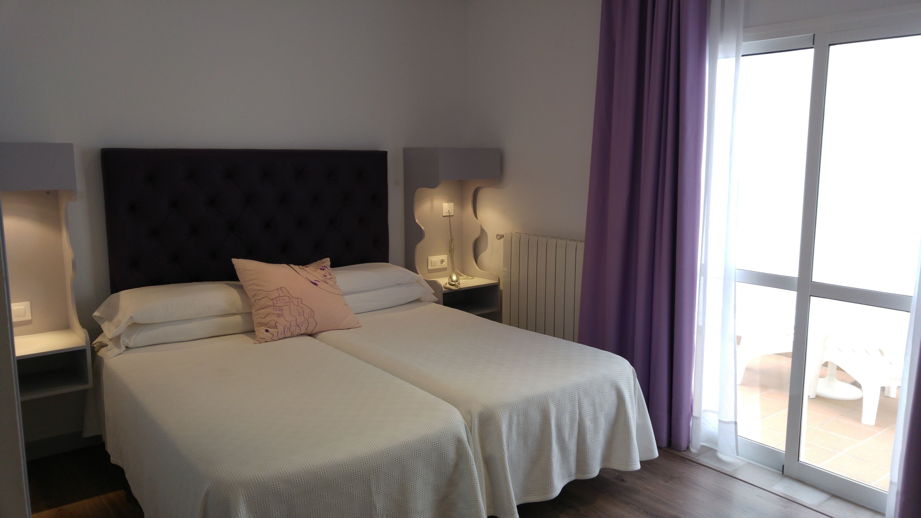 Una cama doble con cabecera, dos mesitas de noche con lámparas, una ventana con cortinas moradas y vistas al exterior.