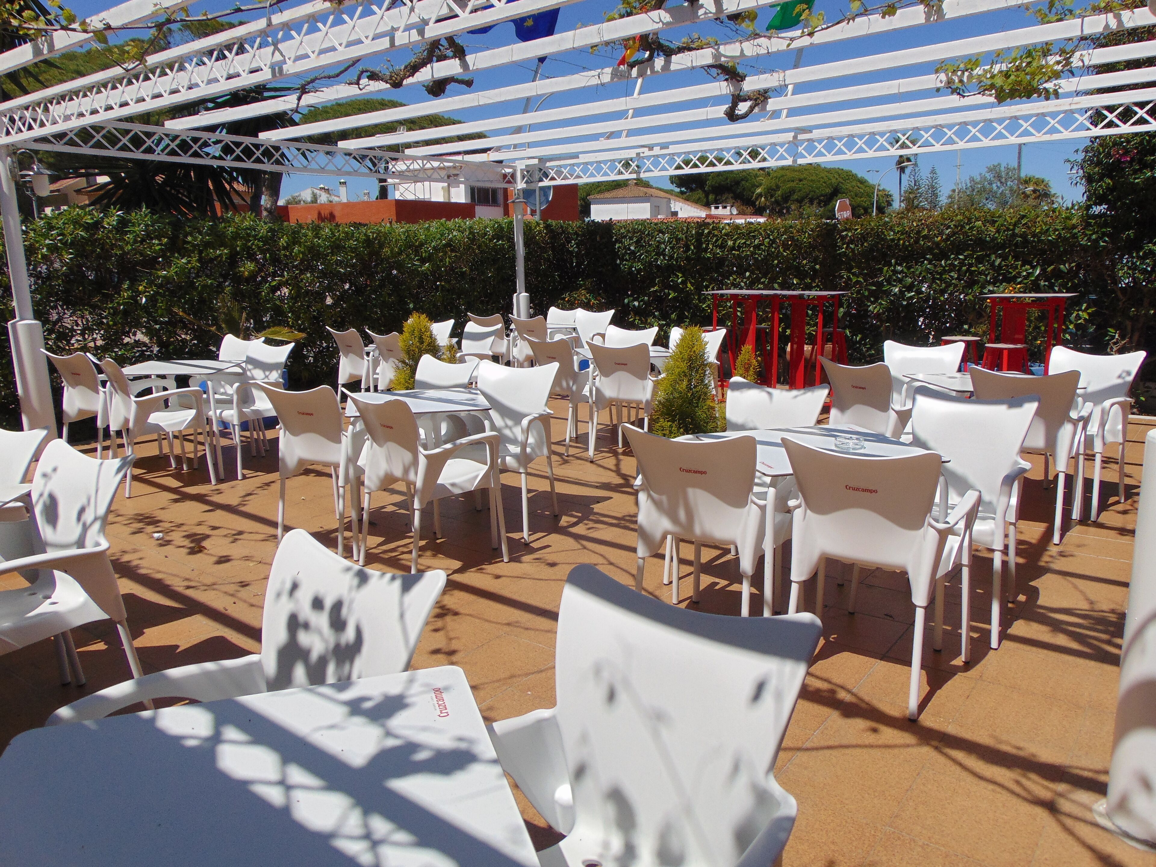 Een terras buiten met witte plastic stoelen en tafels, een witte pergola en een rode tafel op de achtergrond.