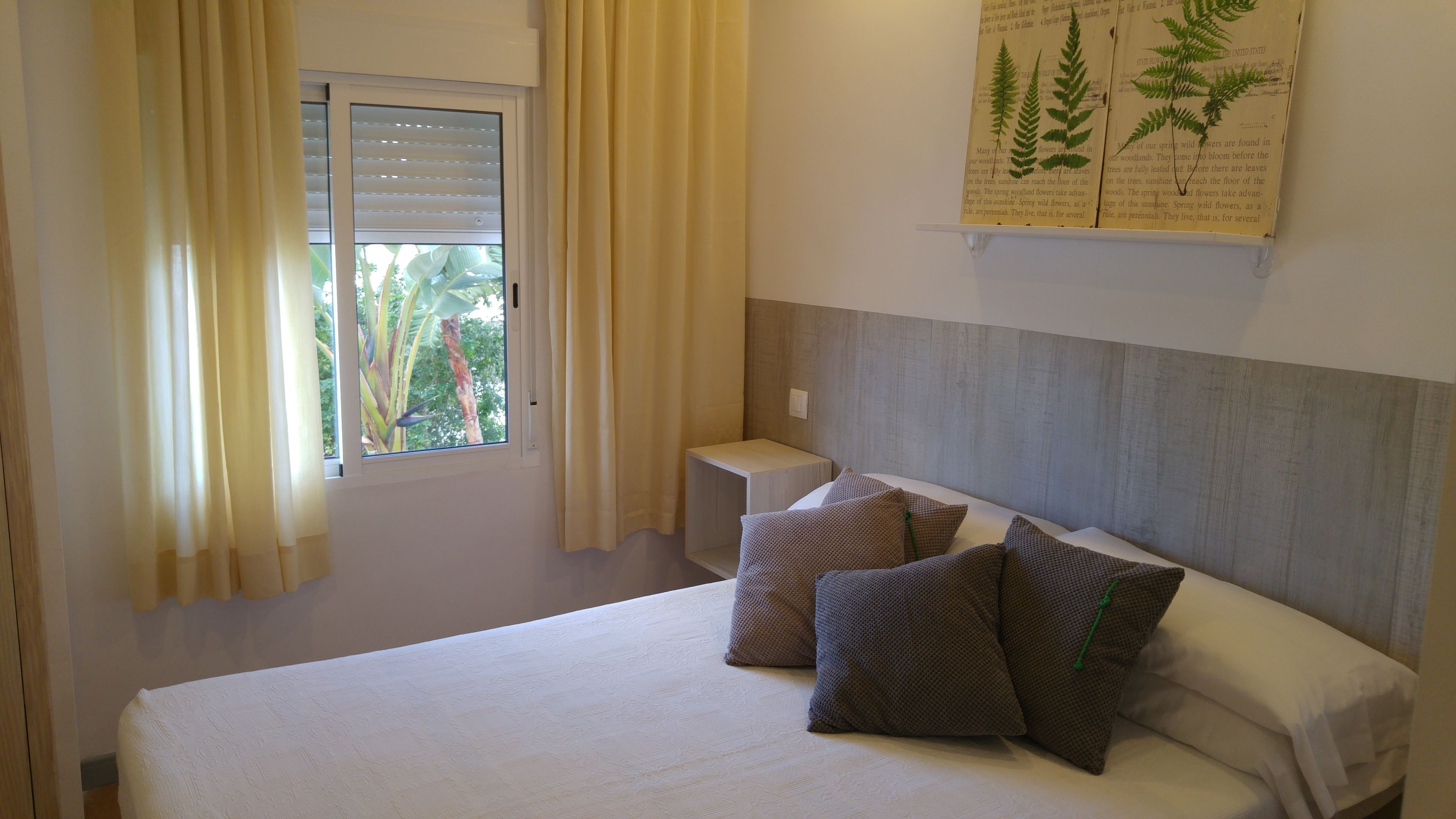 A hotel room with a bed, pillows, a nightstand, a window with curtains, and a wall-mounted botanical illustration.