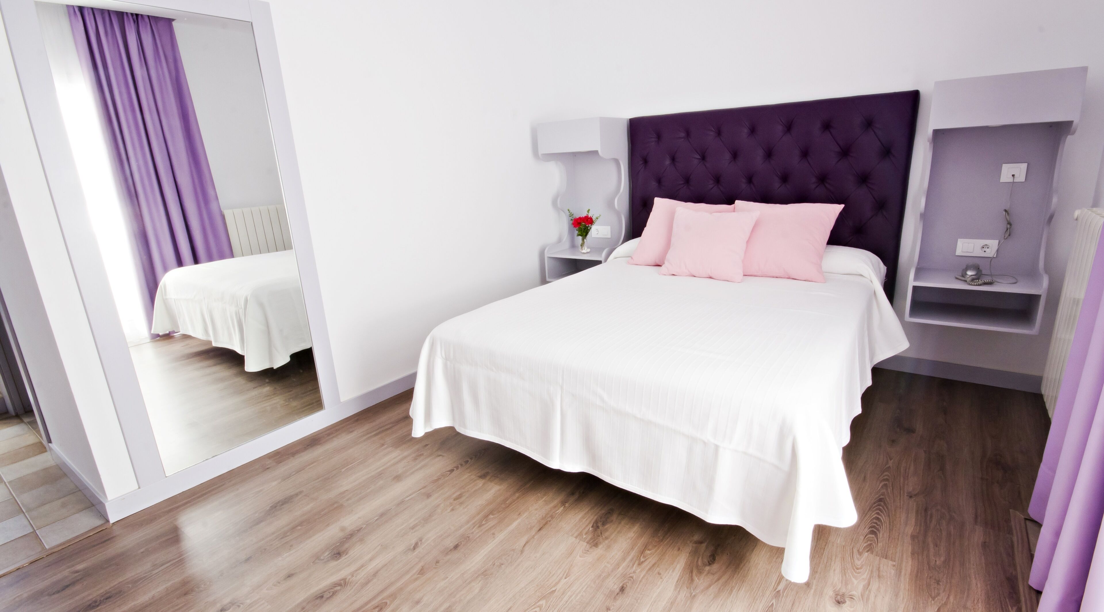 Una habitación de hotel moderna con una cama grande, una mesita de noche, un espejo y un cabecero morado.