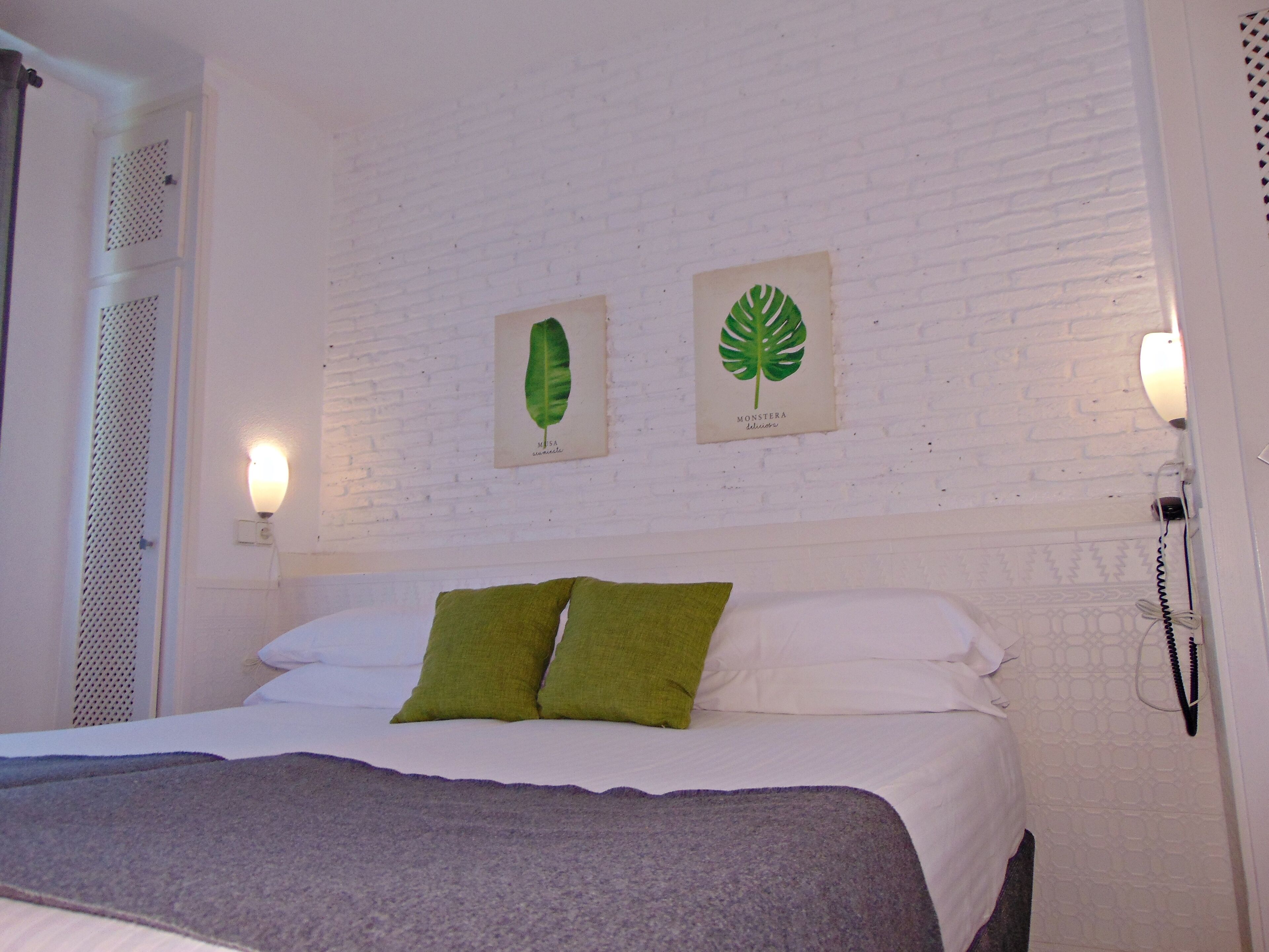 Quarto com cama, almofadas verdes, roupa de cama branca e duas luminárias fixadas na parede.