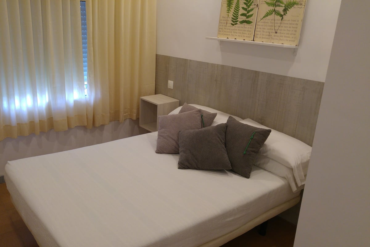 Una cama bien hecha con almohadas, una mesita de noche y una ventana con cortinas.