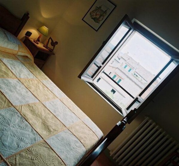 Un dormitorio con cama, una mesita de noche con lámpara, una ventana con vistas a edificios y un cuadro en la pared.