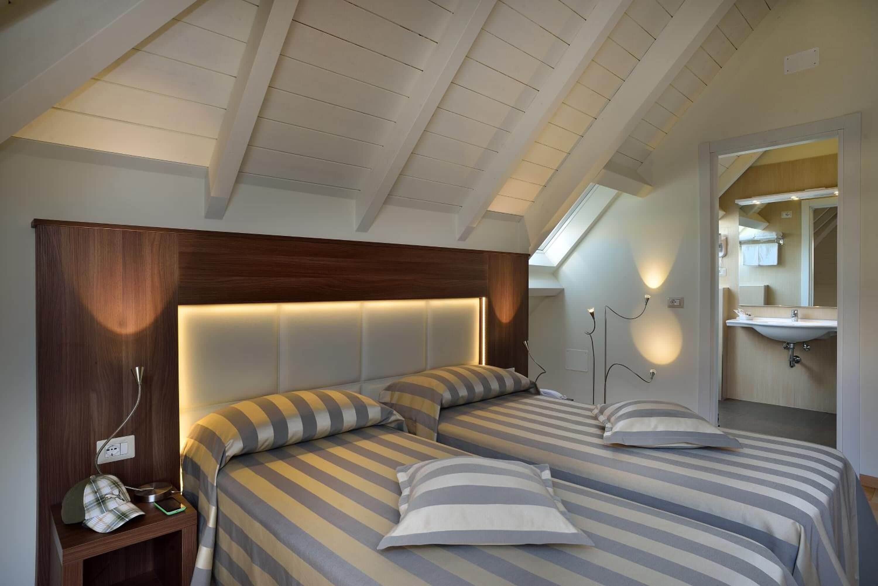 A bedroom with two beds, a wooden headboard, a skylight, a bedside table with a phone, and a bathroom visible through an open door.