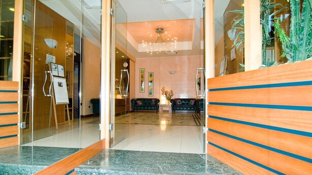 A lobby with a glass partition, a reception desk, a chandelier, and a seating area with a vase of flowers. - Hotel Astor (Bologna)