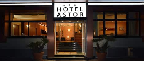 Entrada de un hotel con un letrero que dice 'HOTEL ASTOR', una escalera que conduce al interior y plantas en macetas a ambos lados.