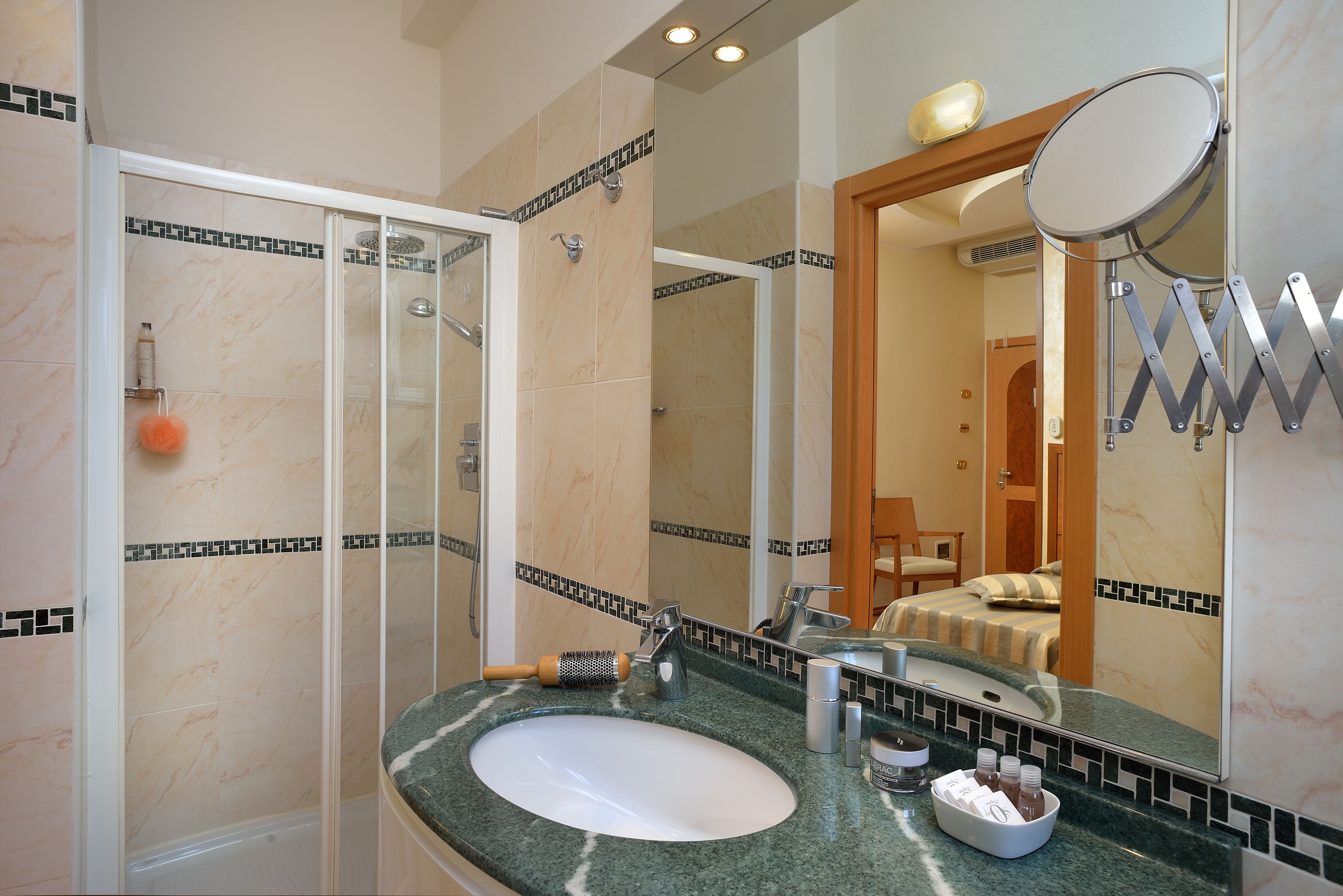 A bathroom with a glass-enclosed shower, a sink with a mirror, and a bed visible through an open door.