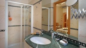 Un bagno con doccia vetrata, un lavabo con specchio e un letto visibile attraverso una porta aperta.