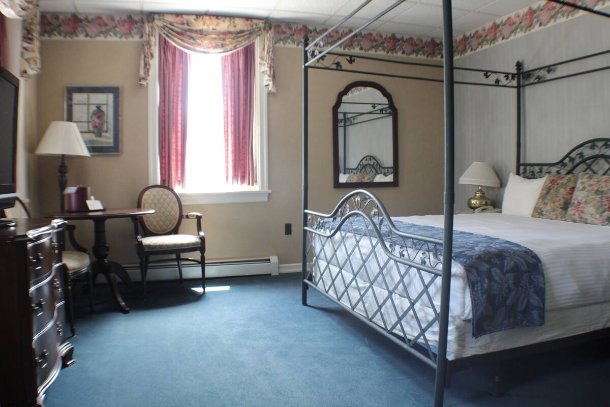 A four-poster bed with a metal frame, a chair, a desk, and a dresser in a room with floral wallpaper and a window with curtains.