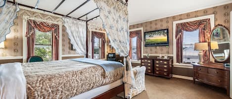 Un letto a baldacchino con struttura in legno e copripiumino floreale, un comò di legno con cassetti, una televisione montata a parete e un'ampia finestra con tende.