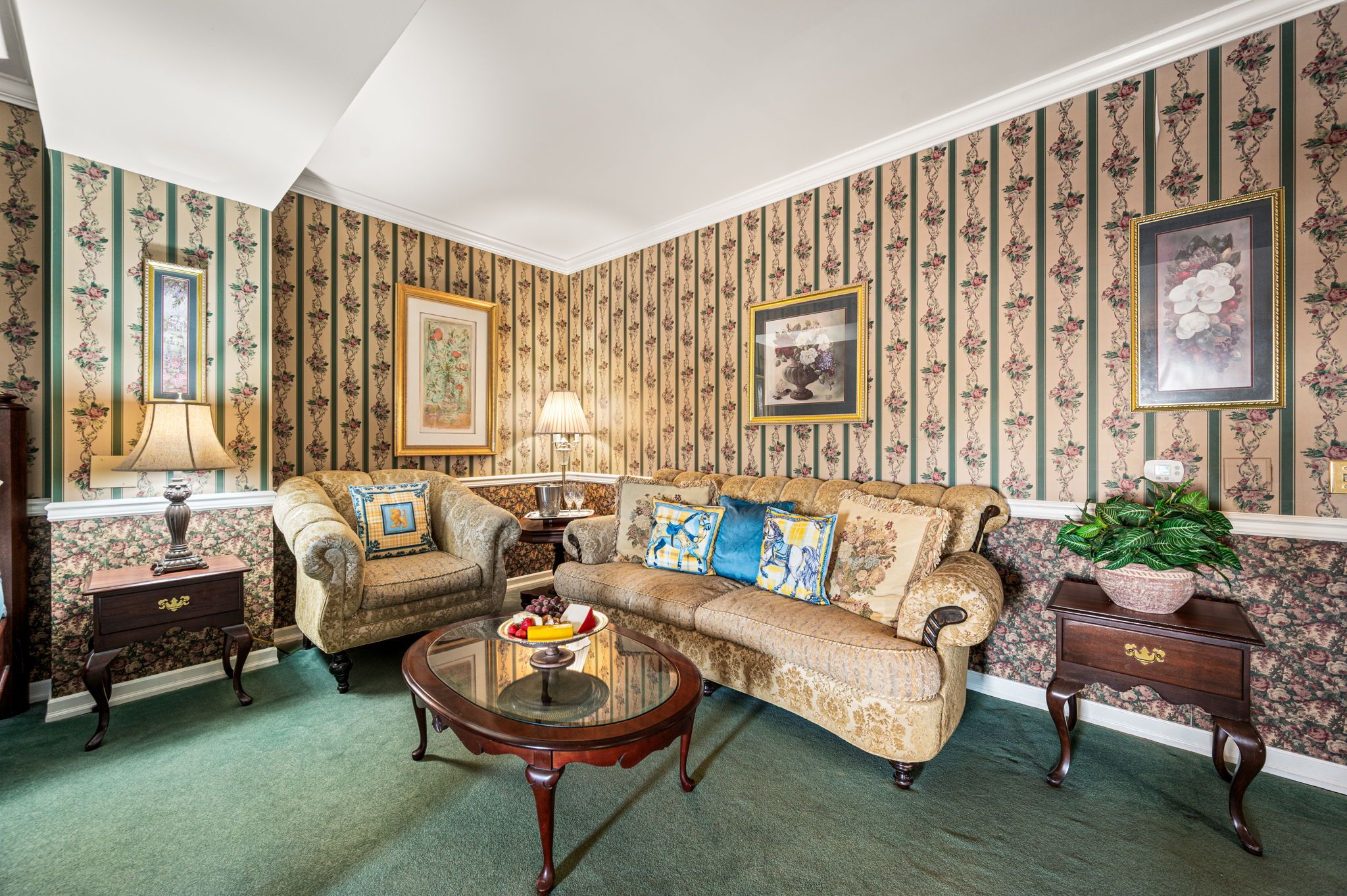 Een klassieke woonkamer met behang met patroon, een zithoek, een glazen salontafel en een bijzettafeltje met een lamp.