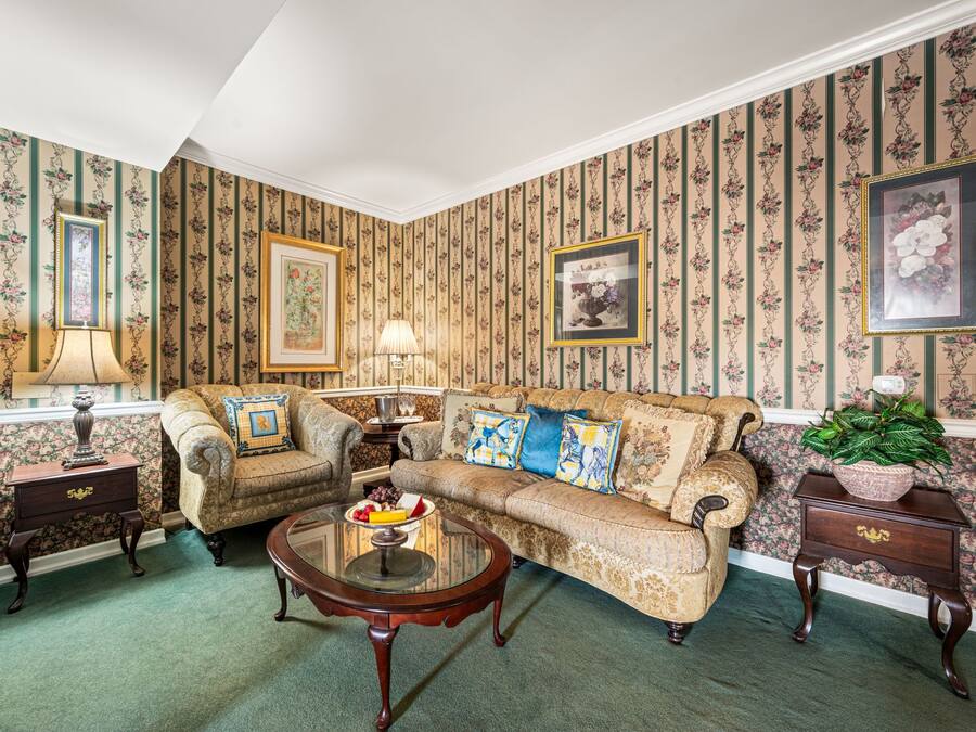 A traditional living room with patterned wallpaper, a sofa set, a glass coffee table, and a side table with a lamp.