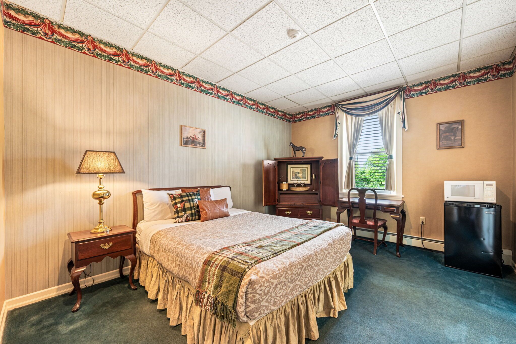A hotel room with a bed, a desk, a chair, a lamp, a picture frame, and a trash bin.