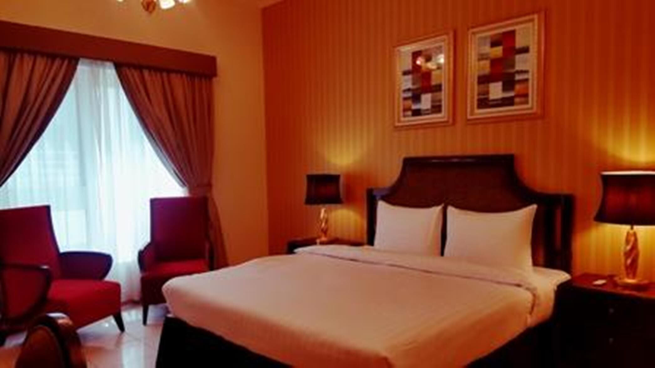 Habitación de hotel con una cama grande, dos mesitas de noche con lámparas, un sillón rojo y una ventana con cortinas.