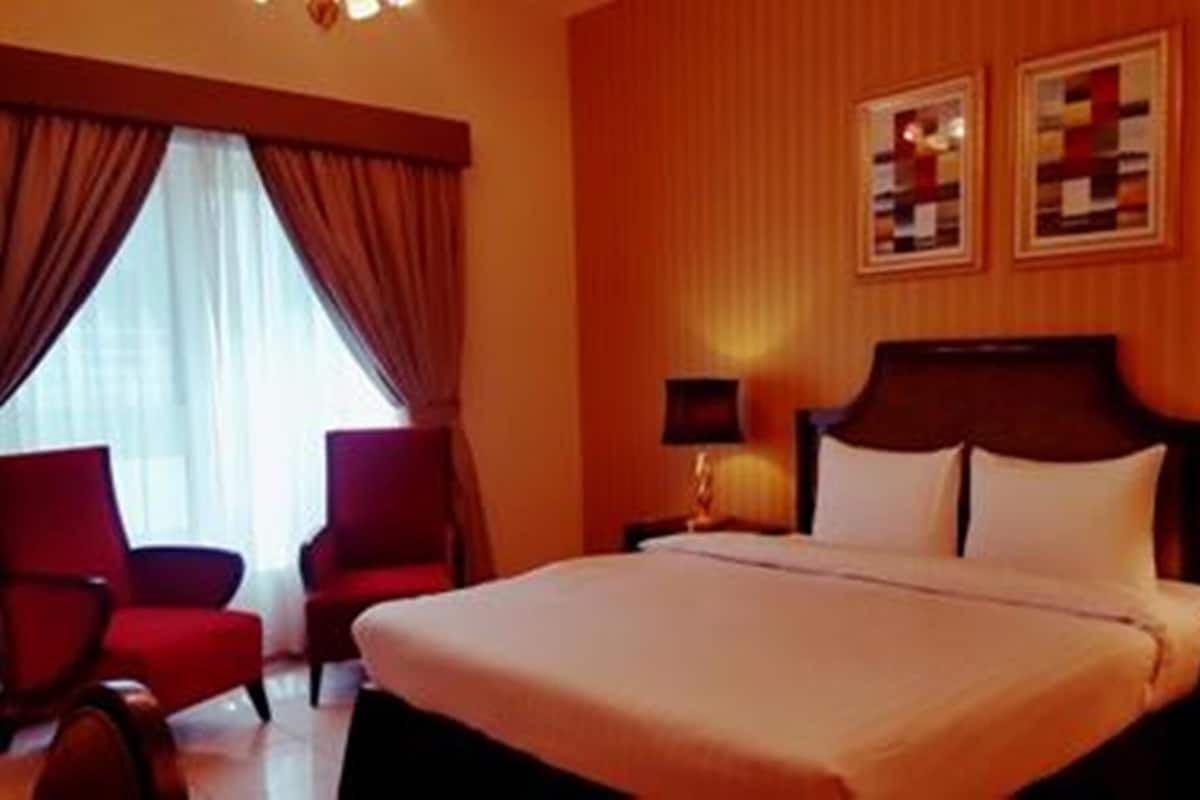 A hotel room with a large bed, two side tables with lamps, a red armchair, and a window with curtains.